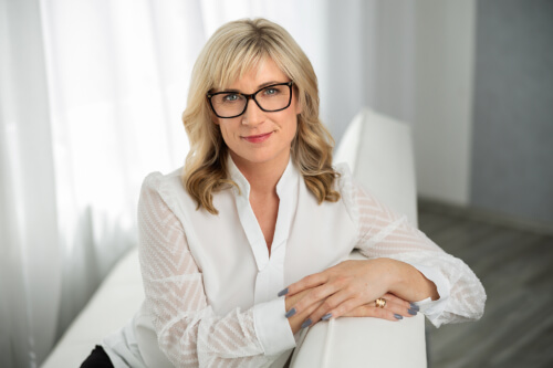 Manuela Tietje sitzt auf einer weißen Couch und lehnt sich darauf. Sie trägt eine Brille und eine weiße Bluse mit durchsichtigen Ärmeln. Ihre Hände sind auf der Lehne gefaltet und sie blickt freundlich in die Kamera. Der Hintergrund ist hell und minimalistisch gestaltet.